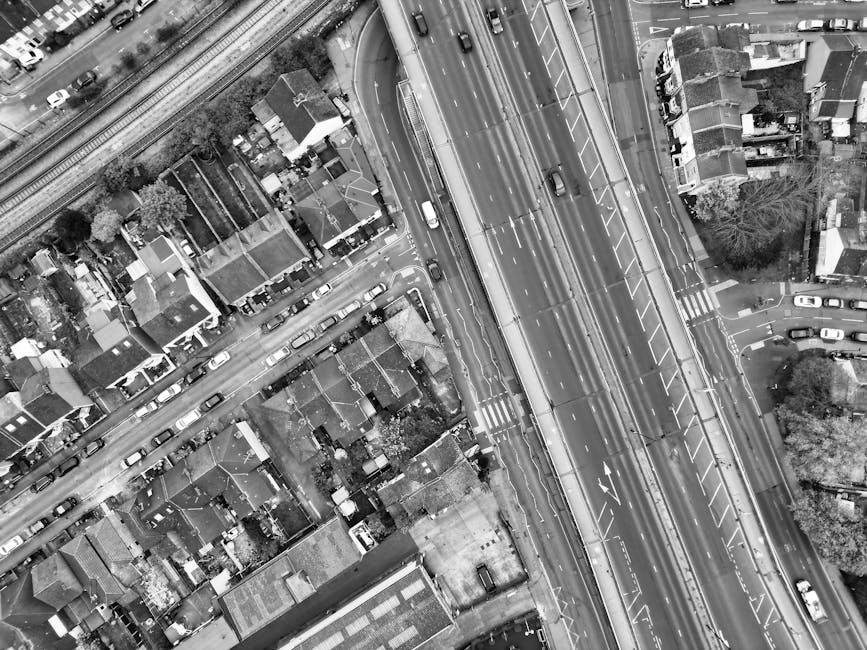 Black and white aerial photograph showing a busy urban scene with a multi-lane road running diagonally across the image, carrying several vehicles including cars and vans. To the left of the road, there are residential houses with small gardens and parked cars along the street. The houses vary in size and design, with some featuring pitched roofs and others flat roofs, and are closely spaced. A railway track runs parallel to the road, located towards the top of the image. On the right side of the road, there is an area of densely packed residential houses and trees. In the foreground, a property with a paved driveway and a gate is visible, with a small vehicle parked outside. The scene captures the typical urban infrastructure involved in house removals, packing, and furniture transport, with [COMPANY_NAME] providing local home relocation and moving services. The photograph emphasizes the logistics of loading and transportation routes necessary for effective house removals from Ilford High Road to the A12 (Seven Kings).
