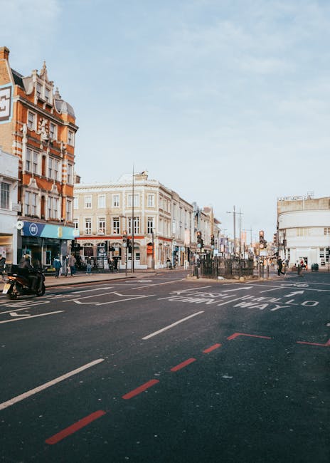 A wide view of a busy street scene on Ilford High Road showing a row of multi-storey buildings with mixed architectural styles, some with ornate brickwork and decorative facades, and others with modern white exteriors. The street has traffic lanes marked with white and red dashed lines, and a section designated for waiting or loading with white text painted on the asphalt. Pedestrians are crossing at the crosswalk and walking along the sidewalk, while vehicles including a black motorcycle and cars are parked or moving along the road. The sky is partially cloudy, providing natural daylight that illuminates the scene. This setting captures the typical environment where home relocation and furniture transport services, such as those offered by Man with Van Seven Kings, operate during professional moving and packing processes in the area.