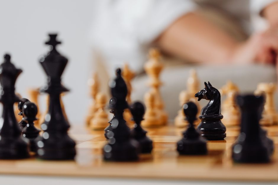 A close-up view of a chessboard during a game, with black and white chess pieces positioned on the wooden surface. The focus is on the black knight in the foreground, with other pieces such as pawns, bishops, and rooks visible but blurred in the background. In the background, a person is partially visible, leaning over the board, suggesting concentration on the game. The setting appears to be indoors with soft lighting illuminating the chess pieces and the person’s arm, which is resting on or near the table. This image captures the intricate details of the chess pieces and the strategic moment in the game, reflecting careful planning and analysis inherent to moving and game setup, similar to the organization involved in house removals or furniture transport. Man with Van Seven Kings may utilize such strategic planning and careful handling in their home relocation services, including packing, moving, and staircase or parking logistics.
