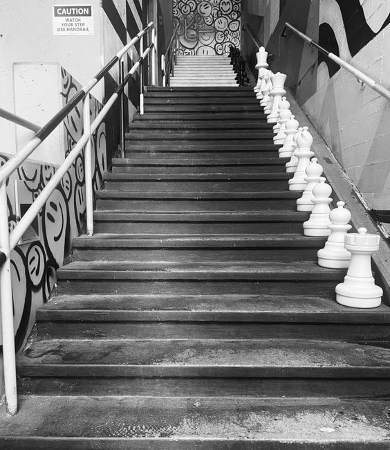 A black and white photograph of a staircase located indoors, with a metal handrail on both sides. Several large white chess pieces, including pawns and a rook, are lined up along the right side of the staircase, positioned on the steps as if ready for a game. The staircase has visible wear on the wooden treads, and the walls beside it are decorated with graffiti-style artwork, creating an urban, artistic environment. At the top of the stairs, a mural with circular patterns is partially visible, adding to the creative atmosphere. This scene is relevant to a house or property relocation context, accentuating the importance of navigating challenging staircases during removals, with [COMPANY_NAME] potentially assisting in the moving process and furniture transport within such environments.