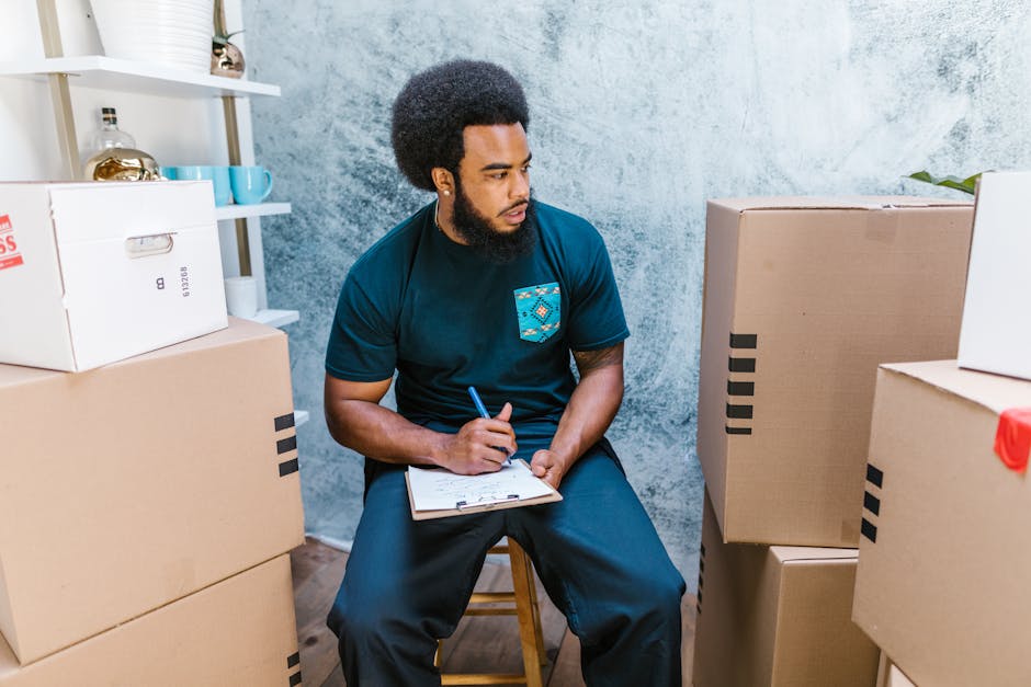 A man with curly black hair and a beard, wearing a navy blue t-shirt with a colourful patterned pocket, is sitting on a wooden stool inside a room with a textured blue wall. He is holding a pen and a clipboard, appearing to be taking notes or making a checklist. Surrounding him are several cardboard boxes of various sizes, some sealed with black tape, and a few filled with packing materials like plastic wrapping. The boxes are arranged on the floor and on shelves, indicating a home relocation or packing process. The environment is well-lit, with natural or artificial light highlighting the boxes and the man's focused expression. This scene reflects the process of packing and planning involved in house removals, supported by a professional moving service like Man with Van Seven Kings, during an ongoing house move or packing task in preparation for furniture transport and home relocation.