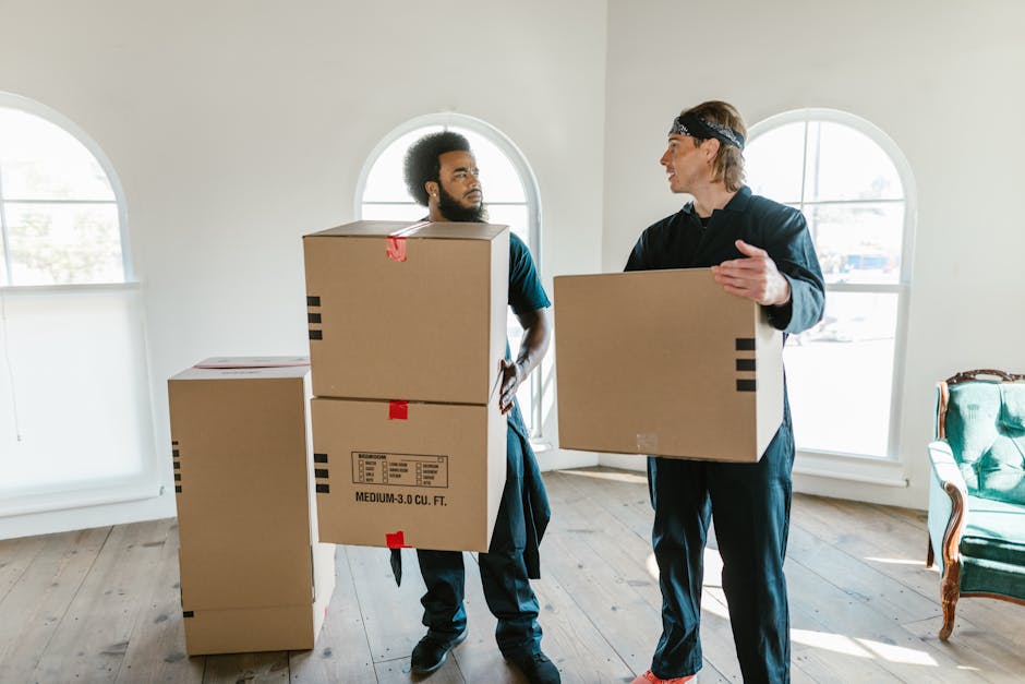 Two movers from Man with Van Seven Kings are inside a well-lit room with white walls and large arched windows, engaged in the packing and moving process for a home relocation. The mover on the left, who has curly hair and a beard, is carrying a medium-sized cardboard box labeled 'MEDIUM-3.0 CU. FT.' and is looking at the other mover. The second mover, with straight hair and a headband, is holding another cardboard box and appears to be explaining or coordinating. Several additional packed boxes are positioned on the wooden floor, some stacked, ready for transport. The room contains minimal furniture, including a green upholstered armchair visible on the right side. The daylight streaming through the windows indicates daytime, and the environment suggests an organized packing and loading process, typical within move-in or move-out scenarios for house removals.