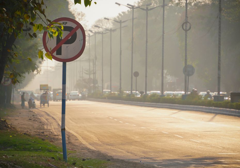 A street scene featuring a no parking sign mounted on a blue and white striped pole in the foreground, with a dusty pavement and a yellow curb edge. The background shows a busy road with lined-up vehicles and some pedestrians on the sidewalk, partially obscured by a light haze or early morning fog. Overhead, there are electrical power lines and streetlights running parallel to the road. Green foliage from trees frames the left side of the image. The scene suggests a typical urban environment, potentially relevant to house removals or furniture transport, with implications for parking regulations and logistical planning for moving services such as those offered by Man with Van Seven Kings.