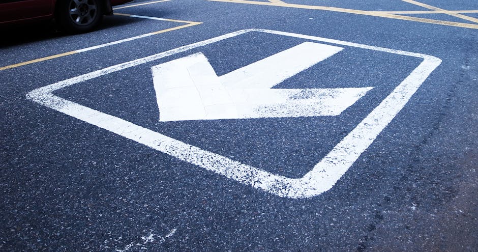 A close-up view of a designated parking area on asphalt pavement, featuring a large white arrow painted within a square outline, indicating direction for vehicle or loading zone usage. The surface shows small gravel and asphalt textures, with clear, bold white markings ensuring visibility. In the background, part of a vehicle’s wheel is visible parked adjacent to the markings, and the area appears to be an outdoor space, possibly near a residential or commercial property. This parking space is relevant for house removals and furniture transport, with the markings assisting in coordinating loading and unloading during a home relocation. The scene highlights the importance of understanding pavement and parking rules, as per local regulations, during the planning and execution of moving services. Man with Van Seven Kings ensures proper parking compliance to facilitate the efficient and safe transport of furniture and packed boxes.