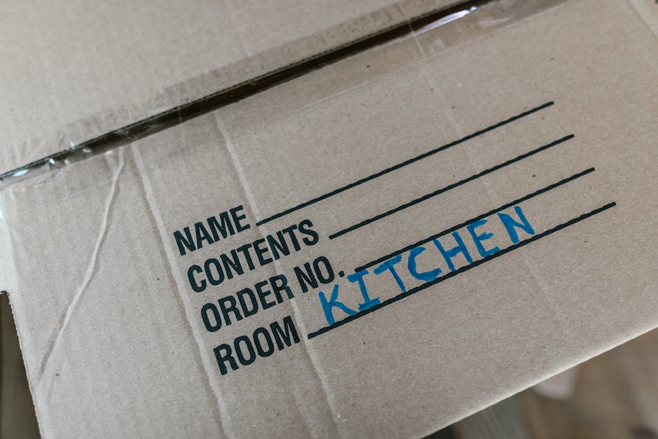 Close-up view of a cardboard moving box with a printed label indicating 'NAME', 'CONTENTS', 'ORDER NO.', and 'ROOM'. The room is marked as 'KITCHEN' in blue handwritten text. The box is sealed with clear packaging tape, and other similar boxes are stacked nearby. The environment appears to be inside a home or storage area during a home relocation process, with the box prepared for furniture transport or packing and moving services. This image was provided by Man with Van Seven Kings, illustrating the packing stage of a house removal in preparation for the transportation of belongings, possibly as part of a professional moving service in SEVEN KINGS.