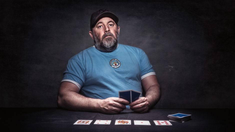 A middle-aged man with a beard, wearing a light blue T-shirt featuring a circular logo on the chest, is seated at a dark table against a black background. He holds a deck of playing cards in both hands, while several other cards are laid face-up on the table in front of him. There is a small stack of additional cards on the right side of the table. The scene is illuminated with focused lighting, highlighting the man’s serious expression and the playing cards, which are a mix of different suits and values. The environment appears to be set up for a card game or magic performance, emphasizing the man’s role in a gaming or entertainment context, with no visible furniture or additional objects related to house removals or moving services. Man with Van Seven Kings occasionally supports the connection to moving and packing processes involved in home relocation and furniture transport, subtly reflected through the arrangement of the objects and the setting's focus on precise handling of the cards.