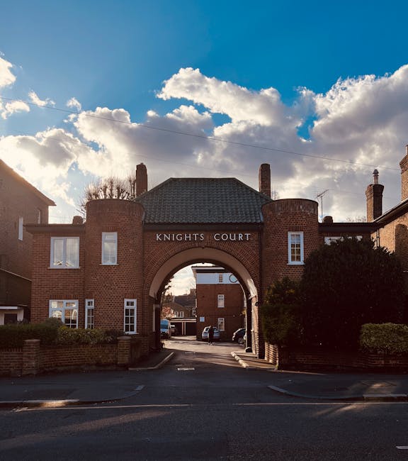 A wide street scene showing the entrance to Knights Court, a brick residential complex with an arched gateway beneath a pitched roof, flanked by two small red brick buildings with multiple white-framed windows. The sky above is partly cloudy with large white clouds and patches of blue, and the sunlight casts shadows on the buildings and pavement. Inside the archway, a paved driveway leads through to parked cars and additional buildings in the background. Surrounding the entrance are well-maintained bushes and low brick walls, indicating a suburban housing area. The scene suggests a typical location for home relocation or moving services, with potential activity related to furniture transport or packing and moving, characteristic of the services provided by Man with Van Seven Kings.
