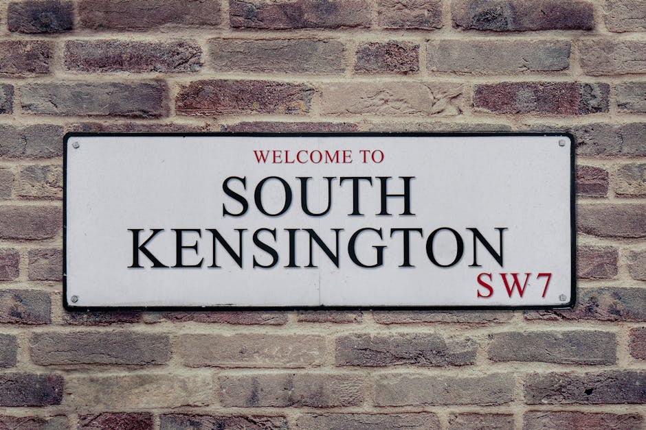 A rectangular street sign mounted on a brick wall reading 'Welcome to South Kensington SW7' with black lettering for the location name and red text for the district code. The sign is positioned at eye level on a textured red and brown brick wall, which provides a neutral background. The sign's surface appears smooth and metallic, with four visible screws securing it to the wall. This scene typically represents an entrance or street identification point in a residential or commercial area. The photograph might be used in content related to house removals or local moving services, emphasizing the importance of precise location identification during relocation processes. The image supports themes of home relocation, specific geographic targeting, and efficient logistics planning for removals companies like Man with Van Seven Kings.