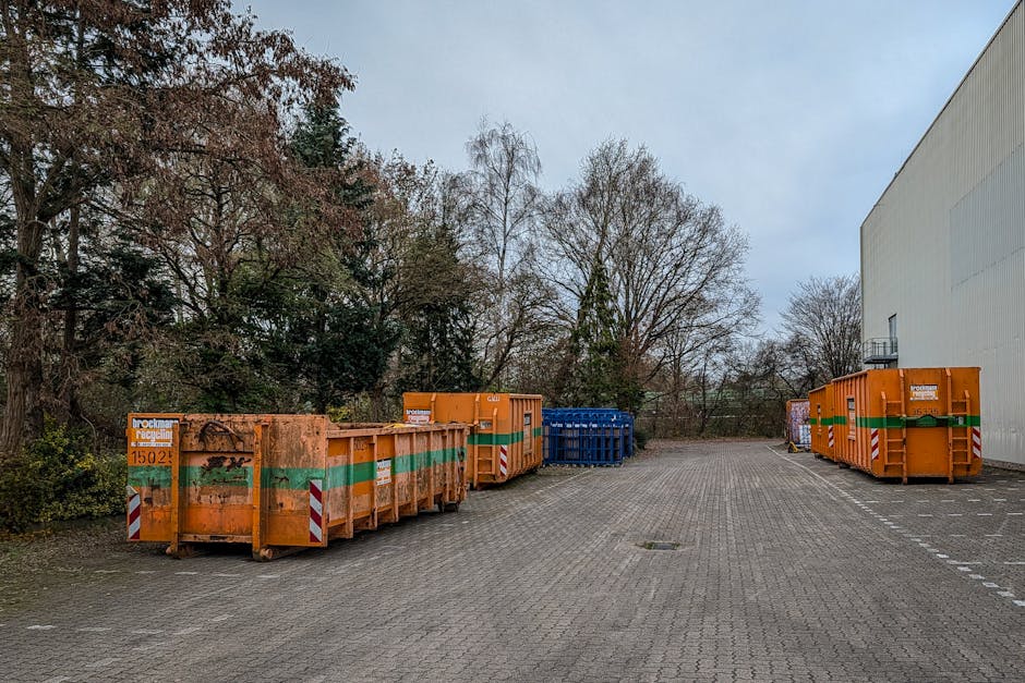 An outdoor paved area adjacent to a large white industrial building features a row of four large orange plastic waste skips with green and white reflective stripes, used for collecting bulky waste mat