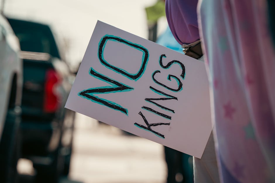 A person holding a white sign with the words 'NO KIDS' written in large, bold, black and blue marker. The individual is standing outdoors, near parked vehicles, with a blurred background that includes cars and a parking lot. The person is partially visible, wearing casual clothing and a purple top, and is seen from behind and to the side, with the sign held in front of their body. This image relates to house removals and moving services, highlighting the importance of clear communication and planning during a home relocation process, as provided by companies like Man with Van Seven Kings. The setting suggests an organized loading or unloading area, where furniture and boxes are moved smoothly to or from a vehicle, with moving equipment or vehicle loading zones possibly nearby.